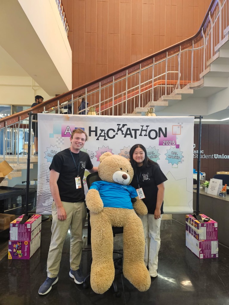 Flora and a teammate with the giant bear in front of the AI Hackathon 2025 banner